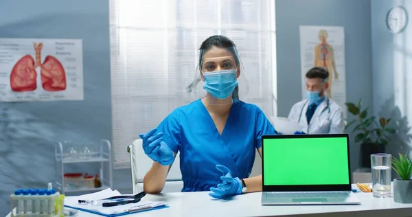 얼굴 가리개를 착용 한 젊은 백인아름다운 간호사의 모습 과 녹색 화면, 말하기, 상담에서 카메라를 보면서 테이블에 앉아 있는 의료 마스크 — 스톡 사진