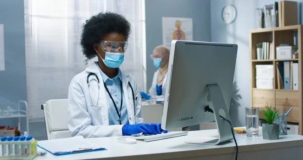 Retrato de una joven afroamericana docotr profesional en máscara médica sentada en el escritorio en el gabinete de trabajo y la búsqueda en línea en la computadora en el laboratorio. Virus Covid-19, hospital, concepto de medicina —  Fotos de Stock