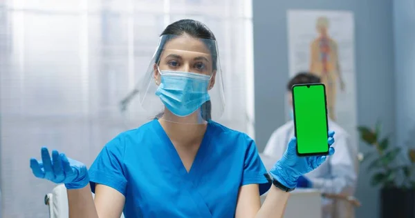 緑の画面で手のスマートフォンで保持保護顔シールドと医療マスクで若い白人可愛いです看護婦の女性の肖像画を閉じます,話して,カメラを見て,医師の相談 — ストック写真