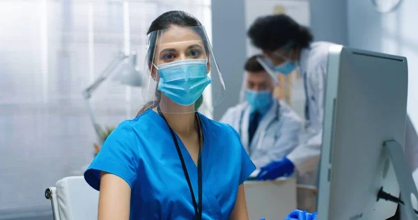 Nahaufnahme Porträt der glücklichen kaukasischen hübschen jungen Krankenschwester mit Maske und Gesichtsschutz, die im Krankenhauslabor arbeitet, während das Coronavirus auf dem Computer surft und in die Kamera schaut und das Konzept der Klinik lächelt — Stockfoto