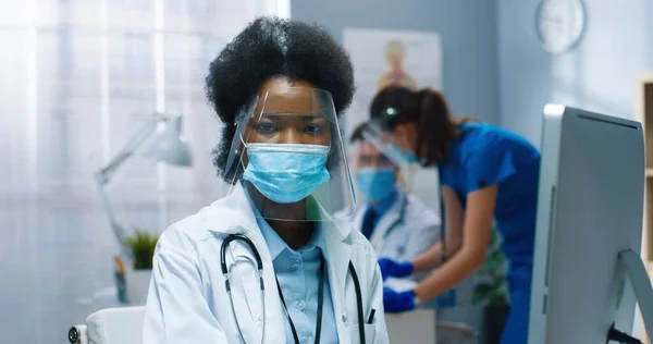 Nahaufnahme Porträt einer glücklichen, hübschen afroamerikanischen Gesundheitshelferin mit Maske und Gesichtsschutz, die im Krankenhauslabor arbeitet, während das Coronavirus auf dem Computer surft, in die Kamera schaut und lächelt — Stockfoto