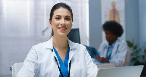 Schöne junge fröhliche positive kaukasische Ärztin in weißem Mantel sitzt im Kabinett in der Klinik bei der Arbeit und schaut in die Kamera und lächelt. Ziemlich glückliche Sanitäterin in guter Laune, Nahaufnahme — Stockfoto