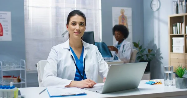 一个快乐快乐的白人年轻美丽的医生的画像，她在橱柜里工作，在笔记本电脑上打字，坐在医院的办公桌前，看着相机，面带微笑。医生的概念 — 图库照片