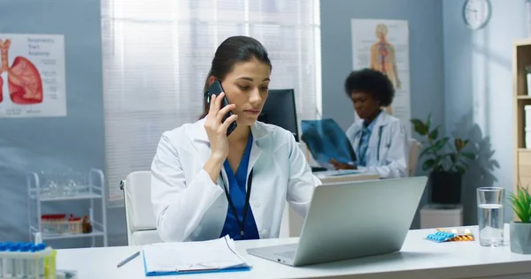 Porträt der kaukasischen jungen schönen Ärztin, die im Kabinett am Schreibtisch im Krankenhaus arbeitet und auf dem Smartphone mit dem Patienten telefoniert. Ärztliches Konzept. Gesundheitsmedizin — Stockfoto