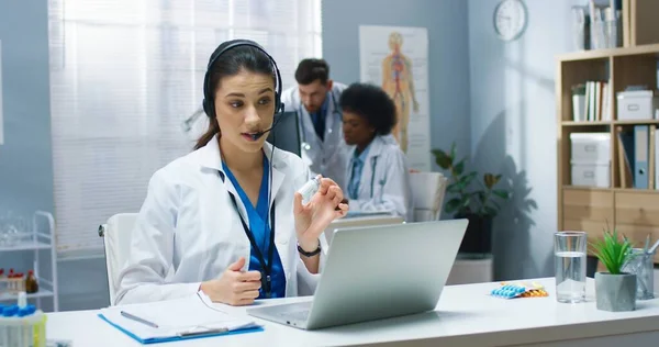 Kaukasische junge hübsche Ärztin mit Online-Videotelefonie-Beratung auf dem Laptop, die im Headset spricht, während die Patientin am Tisch im Krankenhausschrank sitzt. Gesundheitswesen, Porträt, medizinisches Zentrumskonzept — Stockfoto