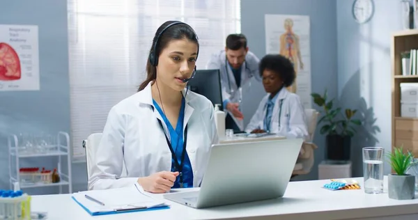 Kaukasische junge hübsche Ärztin mit Online-Videotelefonie-Beratung auf dem Laptop, die im Headset spricht, während die Patientin am Tisch im Krankenhausschrank sitzt. Gesundheitswesen, Porträt, medizinisches Zentrumskonzept — Stockfoto