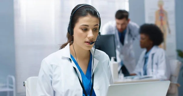 Nahaufnahme einer jungen schönen Ärztin aus dem Kaukasus, die eine Online-Videotelefonie auf einem Laptop hat und mit einem Patienten im Kopfhörer am Arbeitsplatz im Krankenhausschrank sitzt. Gesundheitswesen, Online-Chat — Stockfoto