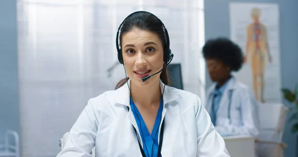 Nahaufnahme Porträt einer hübschen kaukasischen jungen Ärztin in weißem Arztkittel, die mit der Hand in die Kamera winkt und im Kopfhörer im Krankenhauskabinett sitzt. Gesundheitswesen, Videoberatung — Stockfoto