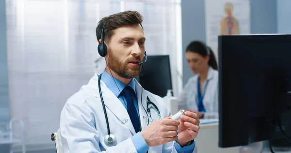 Nahaufnahme Porträt eines jungen, gut aussehenden kaukasischen Arztes, der per Videotelefonie im Headset am Computer im Krankenhauskabinett sitzt und die Impfung zeigt. Medizinische Videokonsultation — Stockfoto