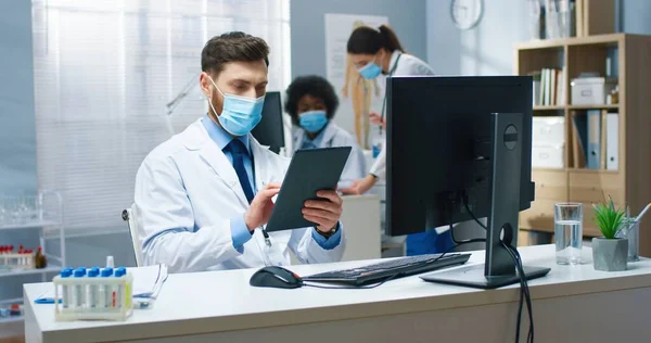 Retrato de bonito branco jovem médico em máscara médica navegando on-line no dispositivo tablet navegar internet trabalhando na clínica no local de trabalho. Conceito do hospital, centro médico, coronavírus — Fotografia de Stock