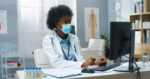 Retrato de un médico profesional afroamericano bastante experimentado en máscara médica y abrigo sentado en el escritorio en el laboratorio de la clínica navegando en el ordenador en el gabinete y mirando a la cámara. Cuarentena del Coronavirus —  Fotos de Stock