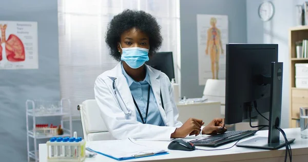 Retrato de un médico profesional afroamericano bastante experimentado en máscara médica y abrigo sentado en el escritorio en el laboratorio de la clínica navegando en el ordenador en el gabinete y mirando a la cámara. Cuarentena del Coronavirus —  Fotos de Stock