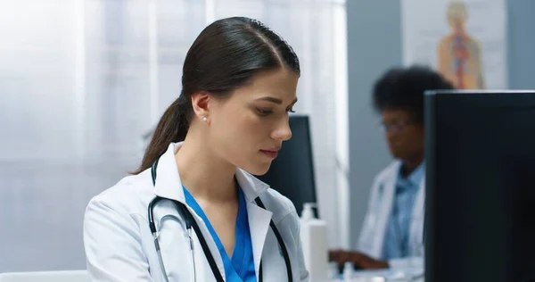 Close up of appy beautiful Caucasian busy young female doctor specialist looking at monitor while typing searching internet on computer working in hospital cabinet. Medical concept, healthcare