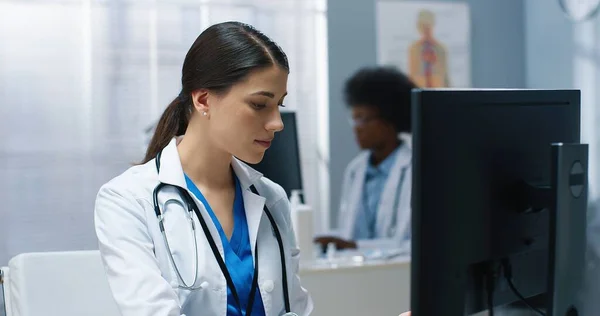 Close up of appy beautiful Caucasian busy young female doctor specialist looking at monitor while typing searching internet on computer working in hospital cabinet. Medical concept, healthcare