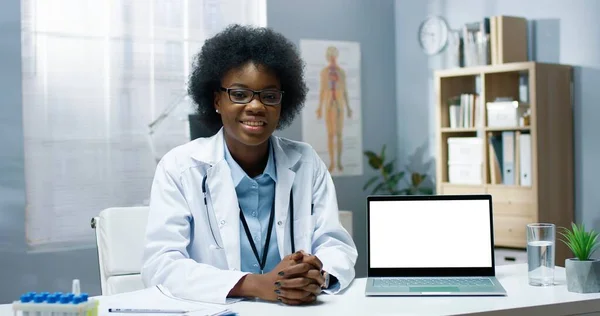 Porträt einer fröhlichen jungen afroamerikanischen Ärztin in weißem Arztkittel, die an einem Tisch im Klinikschrank sitzt und Laptop mit Chroma-Schlüssel zeigt, der im Online-Videochat, Gesundheitswebinar spricht — Stockfoto