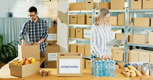 Jovem voluntário caucasiano colocando comida em uma caixa de doação como trabalhador de caridade e membro da comunidade a trabalhar para os pobres. Mulher sênior digitando no laptop em pé no armazém que trabalha na caridade — Fotografia de Stock
