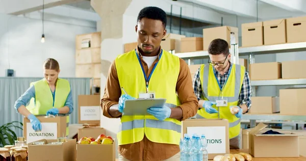 Retrato de feliz jovem afro-americano assistente social masculino em pé na casa de armazenamento verificando lista de doação e sorrindo para a câmera. Voluntários caucasianos embalando alimentos e coisas em caixas conceito de caridade — Fotografia de Stock