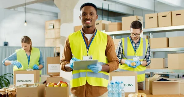 Porträtt av glad ung afroamerikansk manlig socialarbetare som står i magasinet och kollar donationslistan och ler mot kameran. Kaukasiska frivilliga som packar mat och grejer i lådor välgörenhetskoncept — Stockfoto