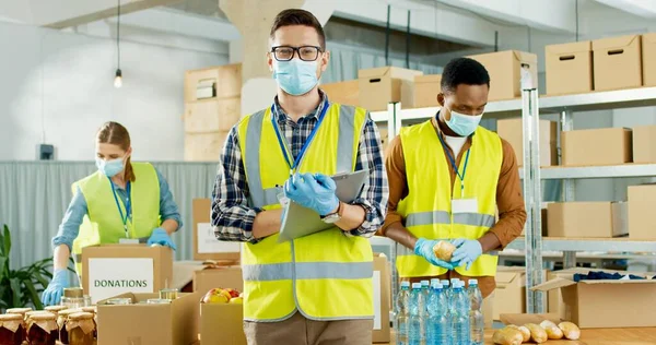 Retrato de jovem caucasiano alegre assistente social em máscara médica em pé no armazém com lista de doação olhando para a câmera. Voluntários de raça mista de uniforme preparando caixas com alimentos para pobres — Fotografia de Stock