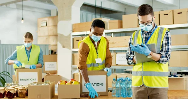 Porträt eines jungen kaukasischen männlichen Managers in medizinischer Maske, der eine Spendenliste schreibt. Mixed-Race Freiwillige arbeiten in Versand Lieferung karitative Lagerorganisation Verpackung Spenden Box — Stockfoto