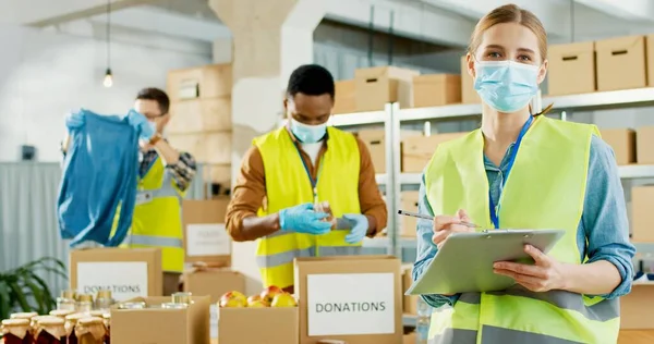 Retrato de jovem caucasiana voluntária feliz em máscara médica e uniforme escrita e sorrindo para a câmera. Trabalhadores de caridade multi-étnicos trabalham na organização de ações caritativas preparando caixa de doações — Fotografia de Stock