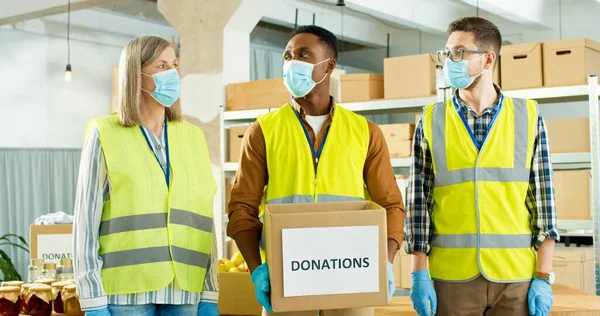Portrait of multi-ethnic happy senior and young male and female charity workers in medical masks in uniform working in donation organization holding donations box for poor. Volunteer concept — Stock Photo, Image