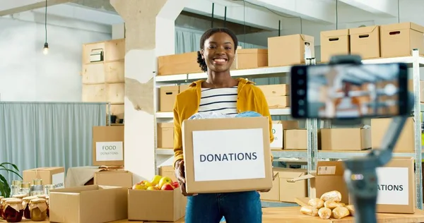 Joven alegre hermosa mujer afroamericana voluntaria de pie en el almacén sosteniendo en las manos caja de donaciones y hablando grabación video blog en el teléfono inteligente. Trabajador social, concepto de trabajo social —  Fotos de Stock