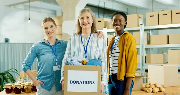 Mezcla de raza feliz maduras y jóvenes trabajadoras voluntarias que trabajan en la entrega de la organización benéfica de stock embalaje caja de donaciones mirando a la cámara y sonriendo, concepto de donación —  Fotos de Stock