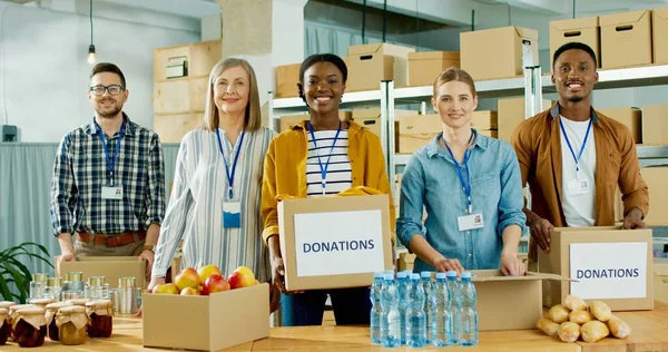 Cheerful young and senior multi-ethnic charity organization workers males and females work at charity warehouse looking at camera and smiling Donation center employees packing parcel volunteer concept