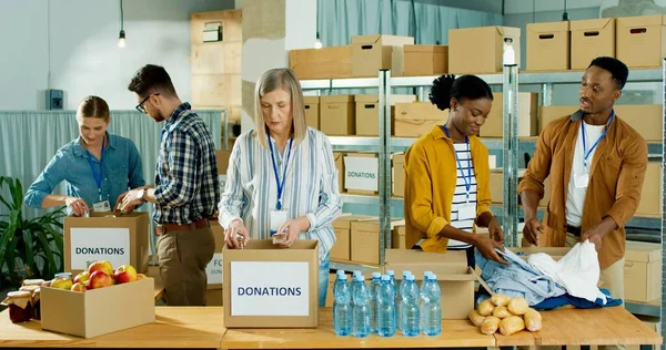 Pessoas caucasianas e afro-americanas homens e mulheres voluntários que trabalham na organização de caridade embalando roupas e mercearia de alimentos em caixa de doações. Voluntariado, ajuda social para pobres e necessitados — Fotografia de Stock
