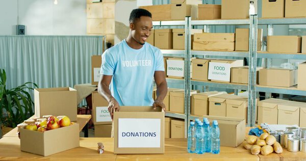 Счастливый афроамериканец, молодой привлекательный мужчина, добровольно улыбается, работая в благотворительном центре, раздавая еду бедным людям. Социальная помощь, пожертвования и волонтерство. социальная работа, гуманитарная помощь