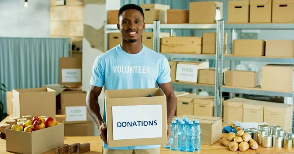 一个年轻的非洲裔美男子的画像，他站在仓库里，手里拿着捐款和社会援助，看着相机，微笑着，慈善中心的概念 — 图库照片