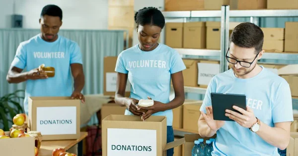 Portrait of Caucasian handsome young guy volunteer tapping on tablet browsing on device making check list of donations working in charity center. African American people packing donation box behind — Stock Photo, Image