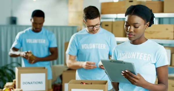 Retrato de una hermosa joven afroamericana concentrada voluntaria de pie en un almacén de organizaciones benéficas escribiendo y revisando donaciones. Trabajadores masculinos clasificando cosas donadas en cajas detrás —  Fotos de Stock