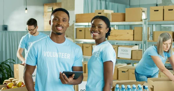 Retrato de alegre casal jovem afro-americano de voluntários masculinos e femininos estão em armazém digitando no dispositivo tablet e sorrindo para a câmera. Assistentes sociais caucasianos empacotam doações em caixas — Fotografia de Stock