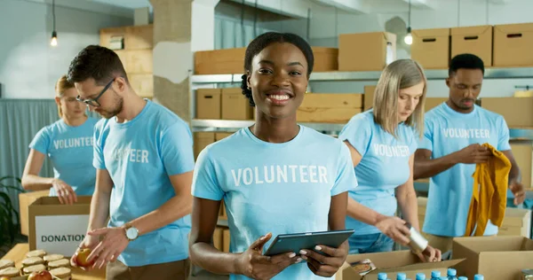 Close up portrait of happy joyful young African American woman in good mood holding tablet in hands and smiling at camera. Charity center volunteer work. Mixed-race volunteers preparing donations — Stock Photo, Image