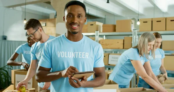 Primer plano retrato de alegre joven afroamericano guapo en el trabajo de buen humor en el centro de caridad, voluntario tocando en la tableta y sonriendo a la cámara. Donaciones, donaciones y voluntariado —  Fotos de Stock