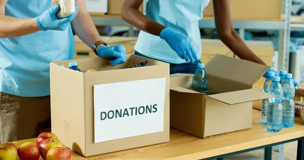 Close up of Caucasian male and African American female hands in gloves packing food and grocery in boxes preparing donation delivery. Charity activity, social help for poor, volunteering and donating — Stock Photo, Image