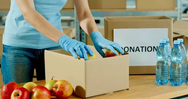 Κοντινό πλάνο του Καυκάσιου εθελοντή χέρια σε προστατευτικά γάντια συσκευασίας τροφίμων σε κουτί δωρεά εργασίας σε φιλανθρωπική τράπεζα τροφίμων. Προετοιμασία δεμάτων δωρεών με μήλα. Πανδημία του κορωναϊού. Δωρεά — Φωτογραφία Αρχείου