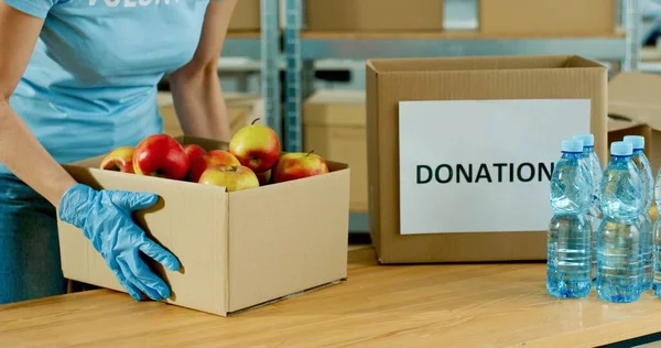 Κοντινό πλάνο του Καυκάσιου εθελοντή χέρια σε προστατευτικά γάντια συσκευασίας τροφίμων σε κουτί δωρεά εργασίας σε φιλανθρωπική τράπεζα τροφίμων. Προετοιμασία δεμάτων δωρεών με μήλα. Πανδημία του κορωναϊού. Δωρεά — Φωτογραφία Αρχείου