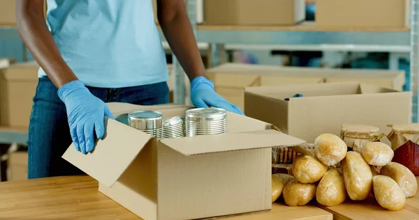 Fechar as mãos de assistentes sociais afro-americanos em luvas de proteção embalando produtos alimentícios em caixa de doação trabalhando em organização de caridade. Doações de alimentos, coronavírus. Doações e voluntariado — Fotografia de Stock