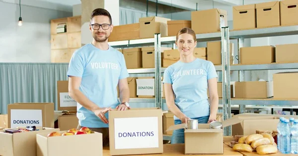 Caucásico joven alegre pareja de hombres y mujeres trabajadores de la caridad poniendo productos alimenticios en la caja de pie en el almacén mirando a la cámara y sonriendo. Los voluntarios preparan donaciones. voluntariado y donación —  Fotos de Stock