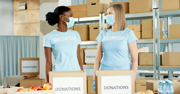 Retrato de mujeres caucásicas y afroamericanas felices de raza mixta trabajadoras sociales en máscaras médicas de pie en entrega de envío donaciones de embalaje de acciones caritativas y mirando el concepto de caridad con cámara —  Fotos de Stock