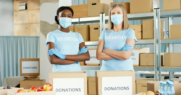 Portrait of positive Caucasian and African American female volunteers in medical masks working in charitable stock organization packing donations sorting food and looking at camera, volunteering — Stock Photo, Image