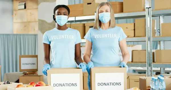 Retrato de mujeres voluntarias positivas caucásicas y afroamericanas en máscaras médicas que trabajan en organizaciones benéficas empacando donaciones clasificando alimentos y mirando cámaras, voluntariado —  Fotos de Stock