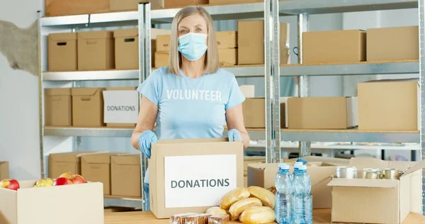 Porträtt av positiv kaukasisk seniormogen kvinnlig volontär i medicinsk mask som arbetar i välgörenhetsorganisation i covid-19 pandemiska förpackningsdonationer sortera mat i låda och titta på kameran — Stockfoto