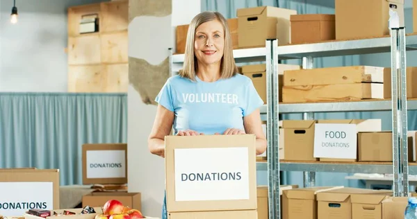Portrait of beautiful Caucasian happy mature woman social worker working in warehouse holding in hands with donations box, looking at camera and smiling. Volunteering and donating, charity concept — Stock Photo, Image