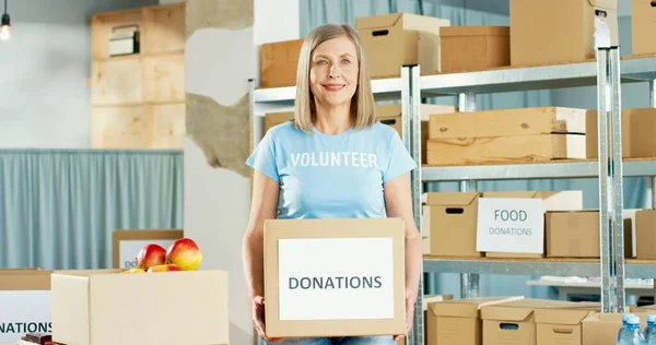 Portrait of beautiful Caucasian happy mature woman social worker working in warehouse holding in hands with donations box, looking at camera and smiling. Volunteering and donating, charity concept — Stock Photo, Image