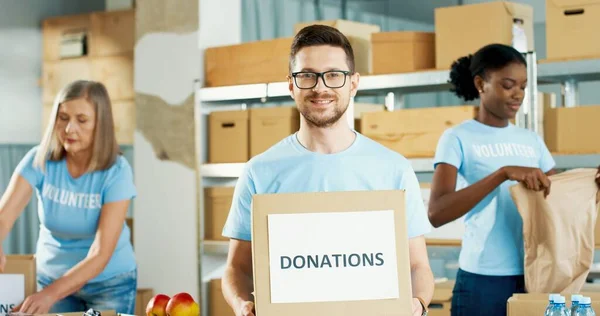 Close up portrait of Caucasian joyful young man social worker stands in warehouse holding in hands with donations box, looking at camera and smiling. Volunteers packing donations on background — Stock Photo, Image