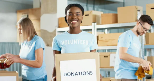 Κοντινό πορτρέτο της Αφροαμερικανής νεαρής ευτυχισμένης γυναίκας εθελοντή που κρατά στα χέρια της κουτί δωρεών, κοιτάζοντας την κάμερα και χαμογελώντας. Φιλανθρωπική οργάνωση. Συνάδελφοι συσκευασίας δωρεές στο παρασκήνιο — Φωτογραφία Αρχείου